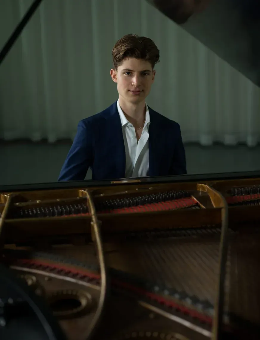 Pianodocent Maxim Hijmerink uit Amsterdam in een donkere blazer staat achter een open vleugel in een zacht verlichte kamer.
