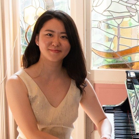 Pianodocent Misaki Yamada in een crèmekleurige jurk zit bij een piano in een lichte kamer met glas-in-loodramen.