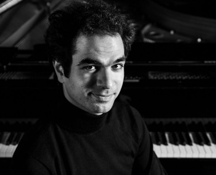Pianodocent Orestes Dimitriadis uit Utrecht  in een coltrui naast een piano, die in de camera kijkt.