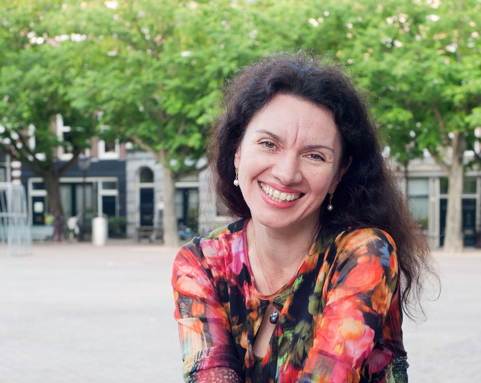 Pianodocent Tatiana Chevtchouk in een kleurrijke blouse buiten, met bomen en een gebouw op de achtergrond.