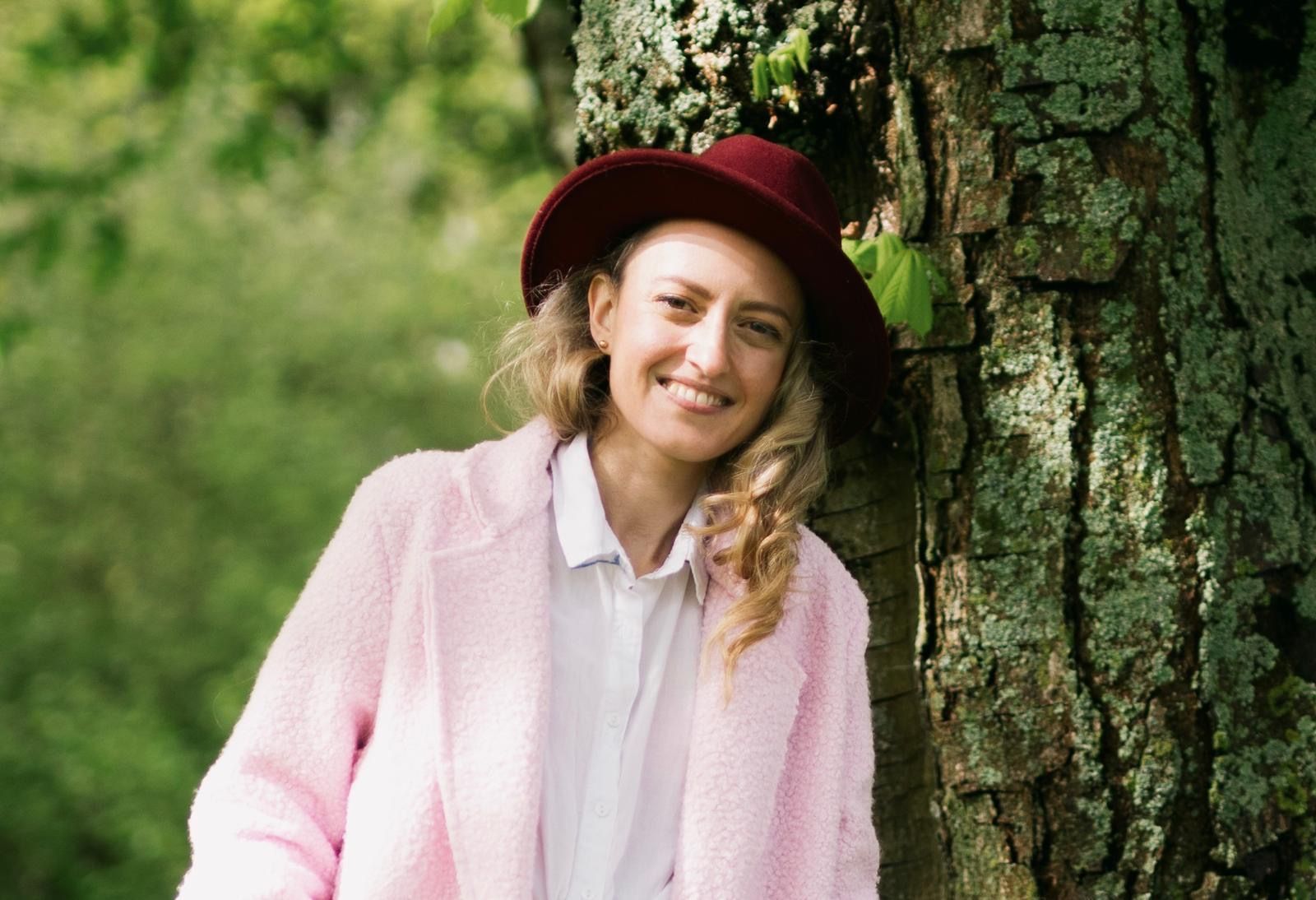 Pianodocent Viktoria Wilde met een rode hoed en een roze vest leunt buiten tegen een boom.