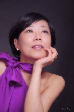 Pianodocent Yumi Toyama  in een paarse sjaal kijkt omhoog, haar kin rustend op haar hand, tegen een donkere achtergrond.