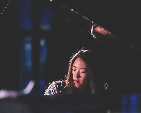 Pianodocent Yunah Han  uit Amsterdam met gesloten ogen onder gedempt blauw en roze podiumlicht, met een hand boven haar hoofd.