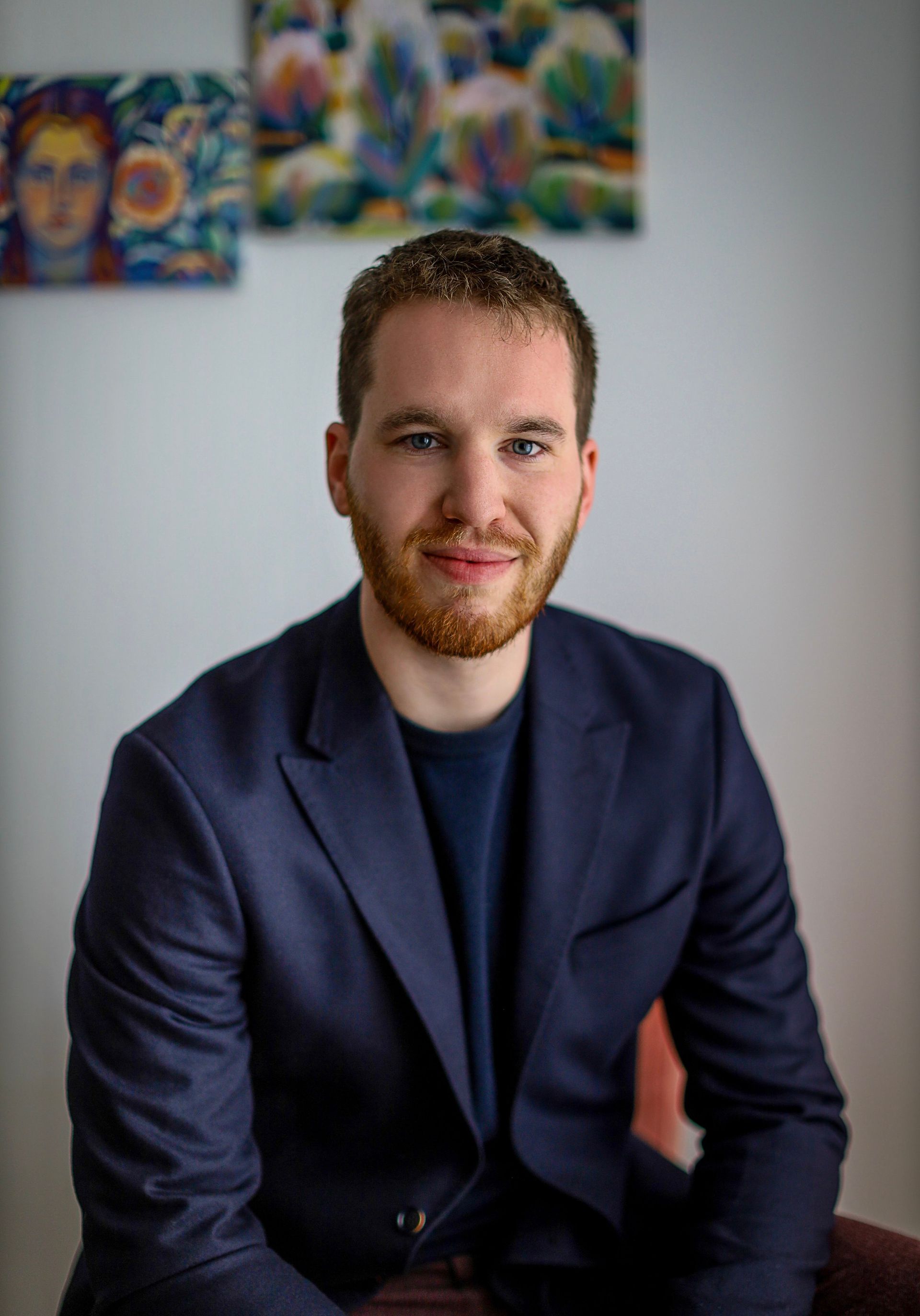 Pianodocent Aron Gunst  in een donkerblauwe blazer zit binnen en glimlacht tegen een lichte muur met kleurrijke kunstwerken.