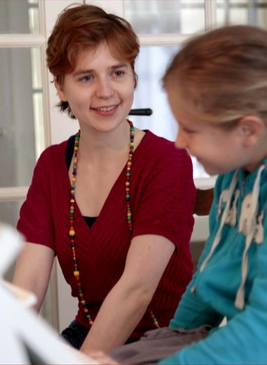 Pianodocent Jana Neplechovitsj met haar pianoleerling  praten binnenshuis, de ene in een rode top en met een kralenketting, de andere in een blauwe hoodie.
