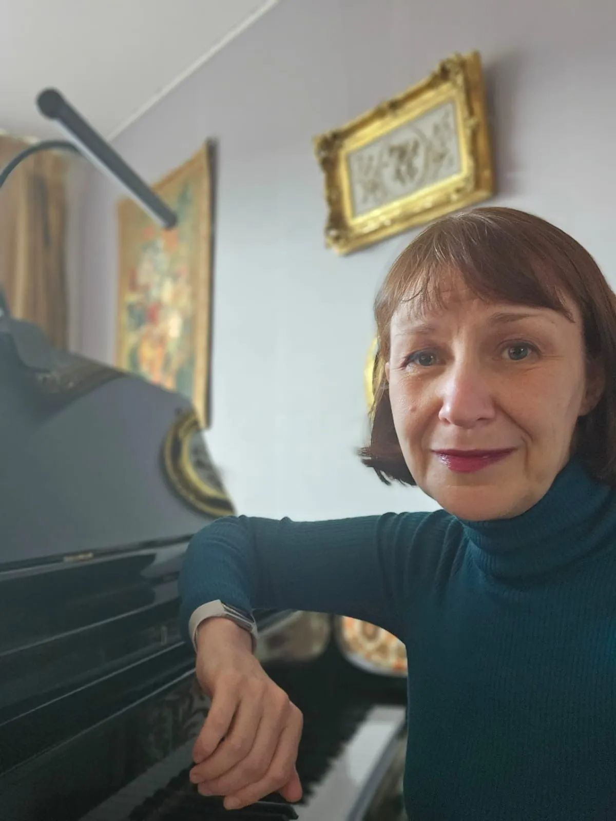 Pianodocent Marina Glotzbach leunt tegen een zwarte piano in een kamer met kunstwerken in gouden lijsten en kijkt in de camera.