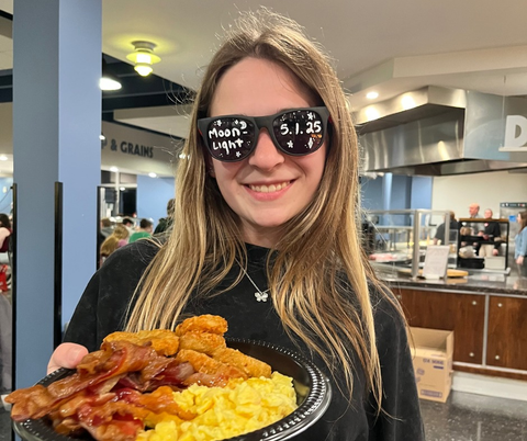 A person smiling while wearing sunglasses holds a bowl of food.