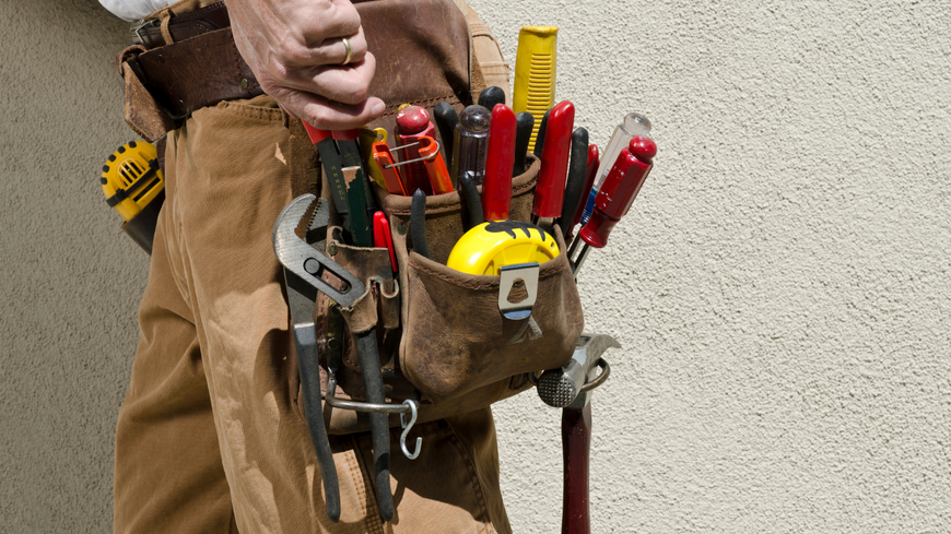 Tool belt with pliers, screwdrivers, tape measure, and wrench worn by a worker against a beige wall