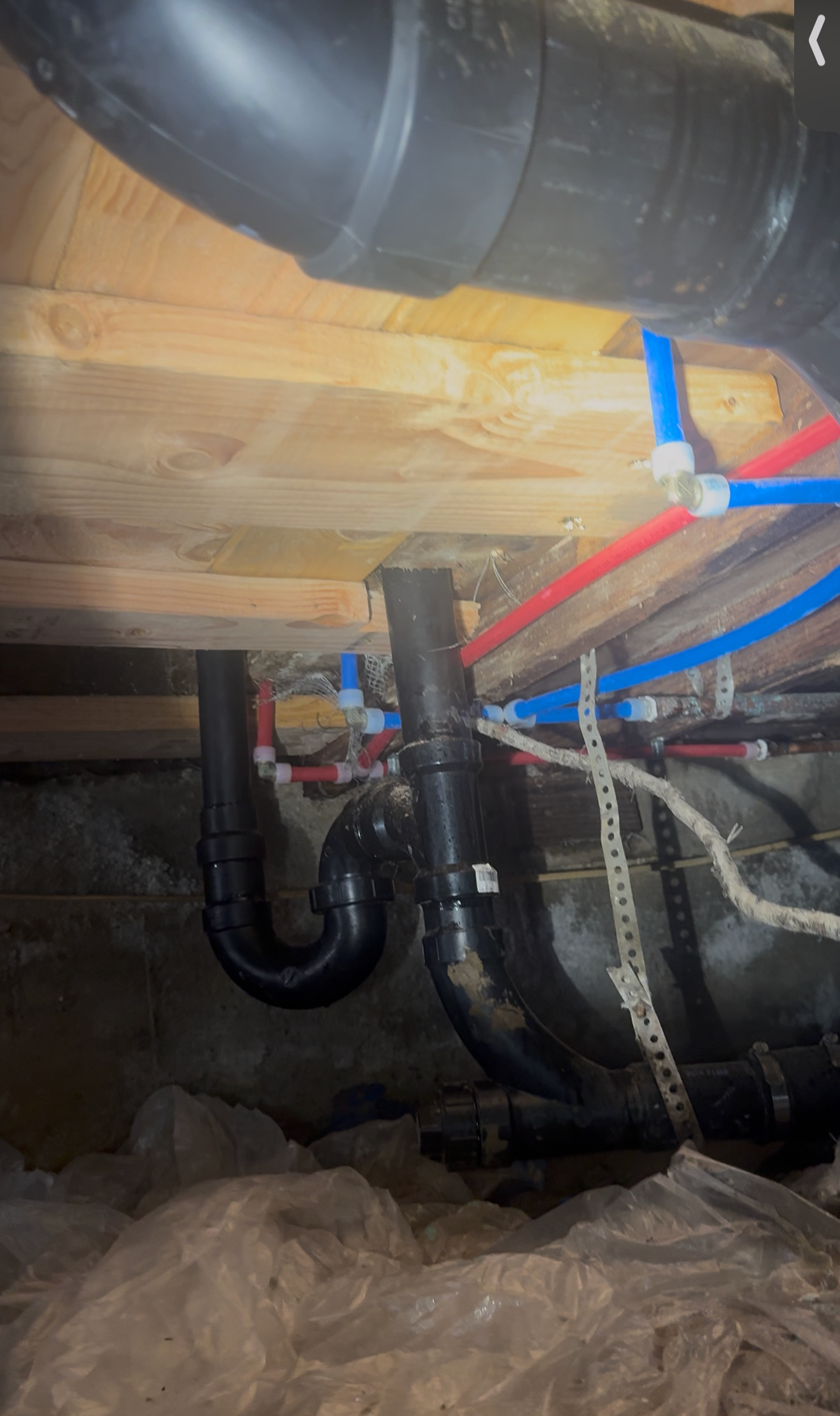Under-floor plumbing with black drain pipes, red and blue water lines, and insulation.