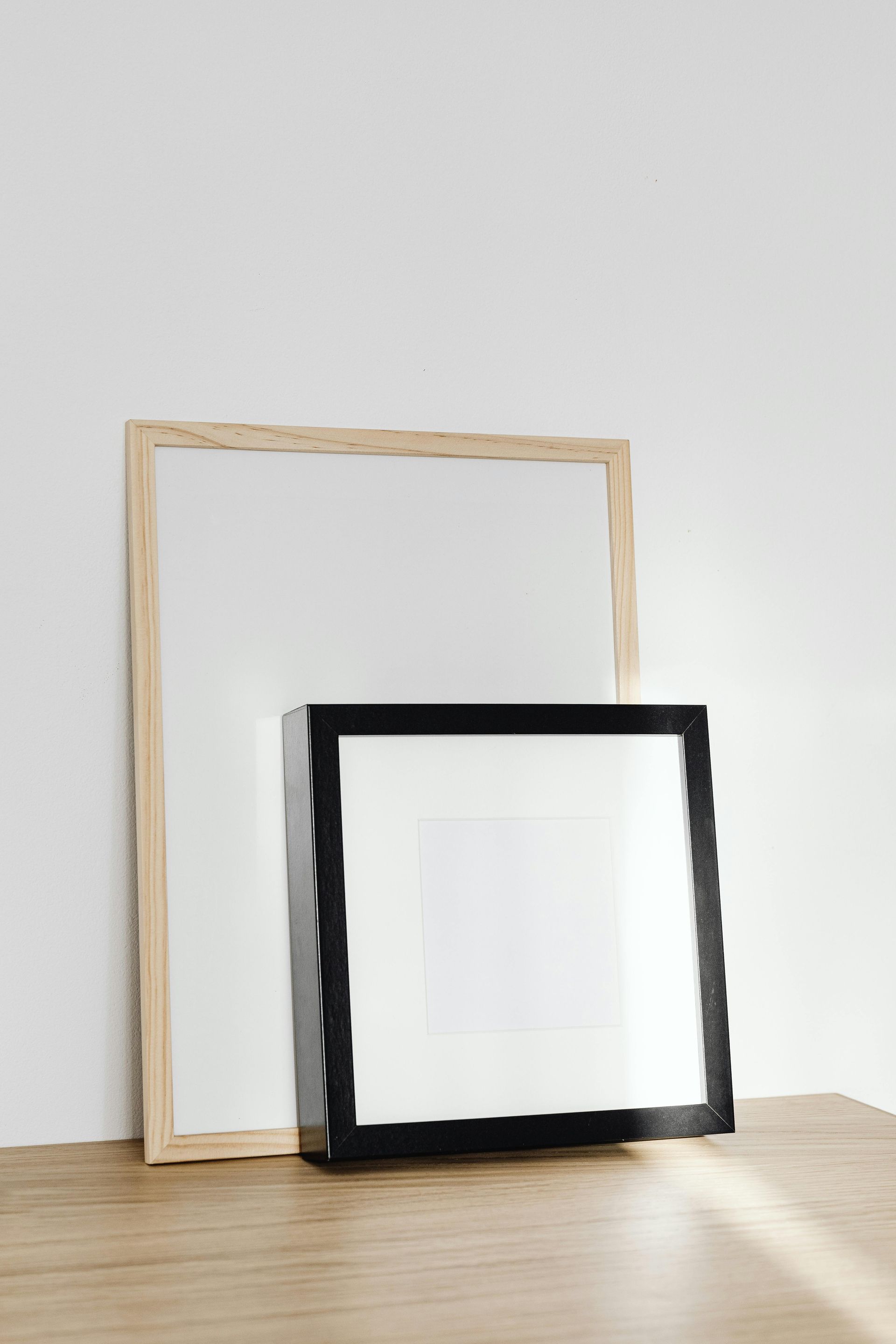 Two empty frames, one light wood and one black, leaning against a wall on a wooden floor.