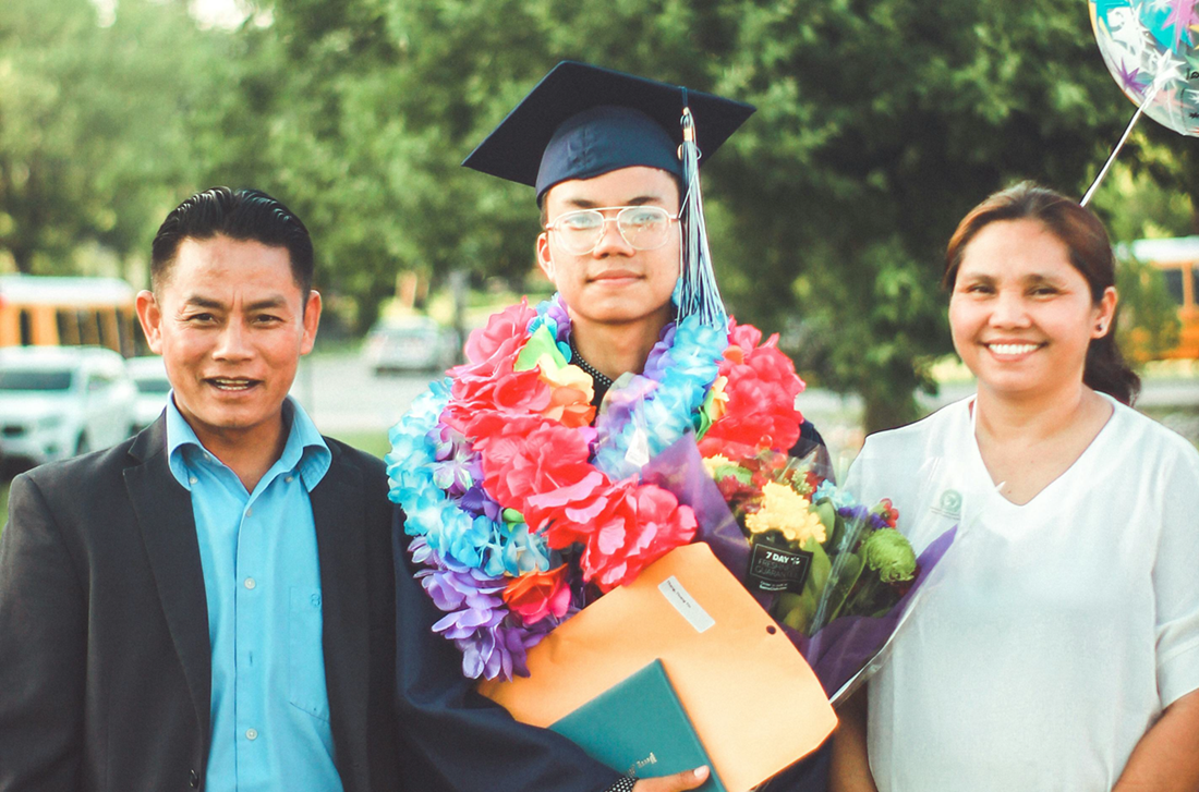 Graduate in cap and colorful lei holding a diploma, standing between two smiling people outdoors.