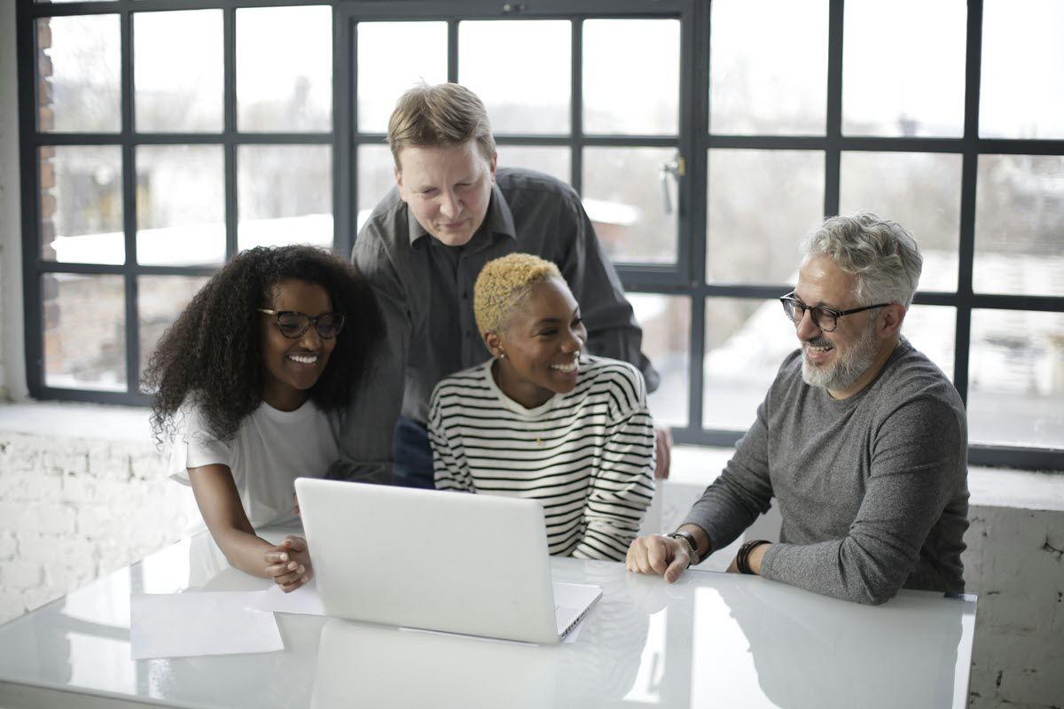 Four people gathered around a laptop in a bright office, collaborating and smiling.