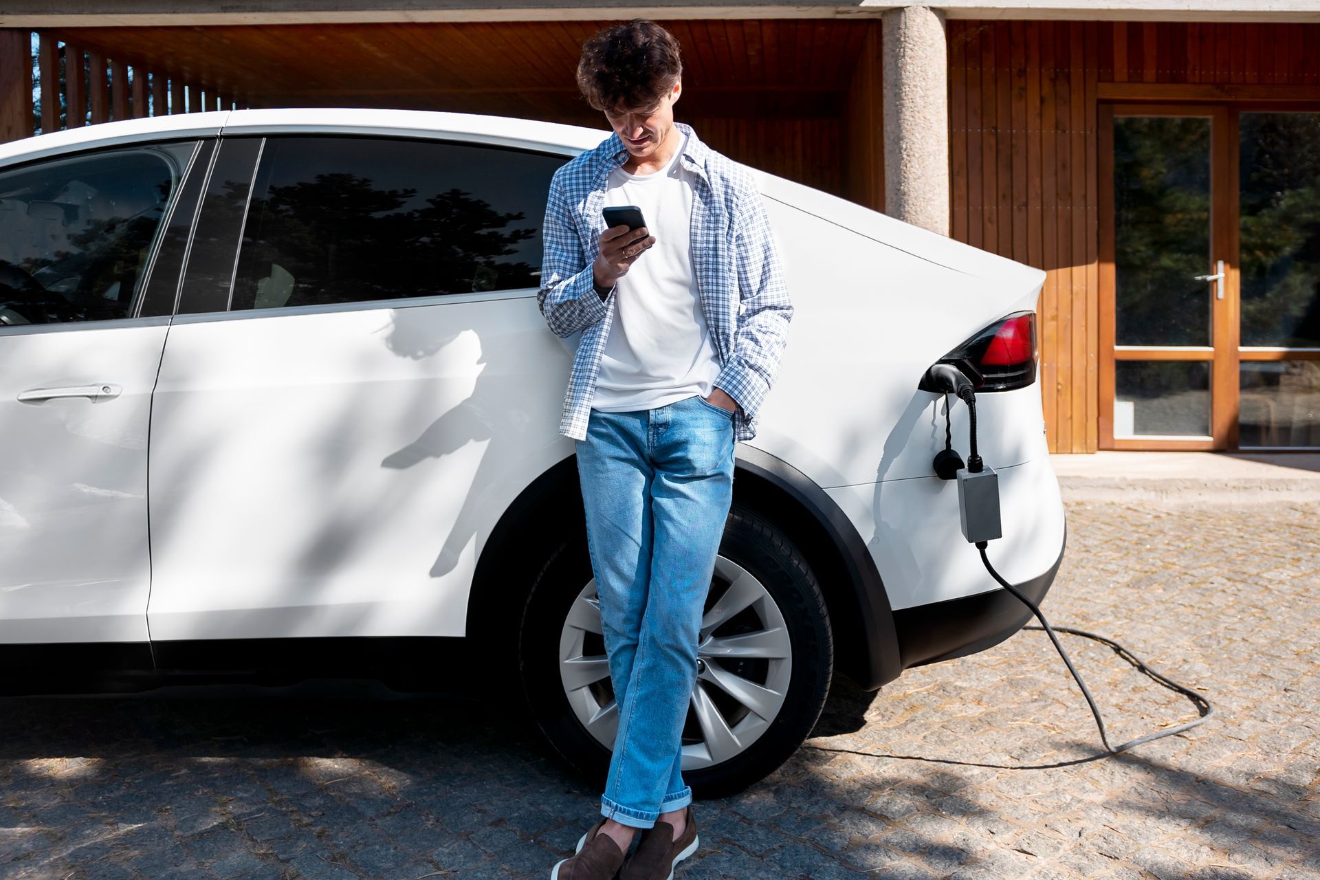 Person charging a white electric SUV while looking at a phone in a driveway