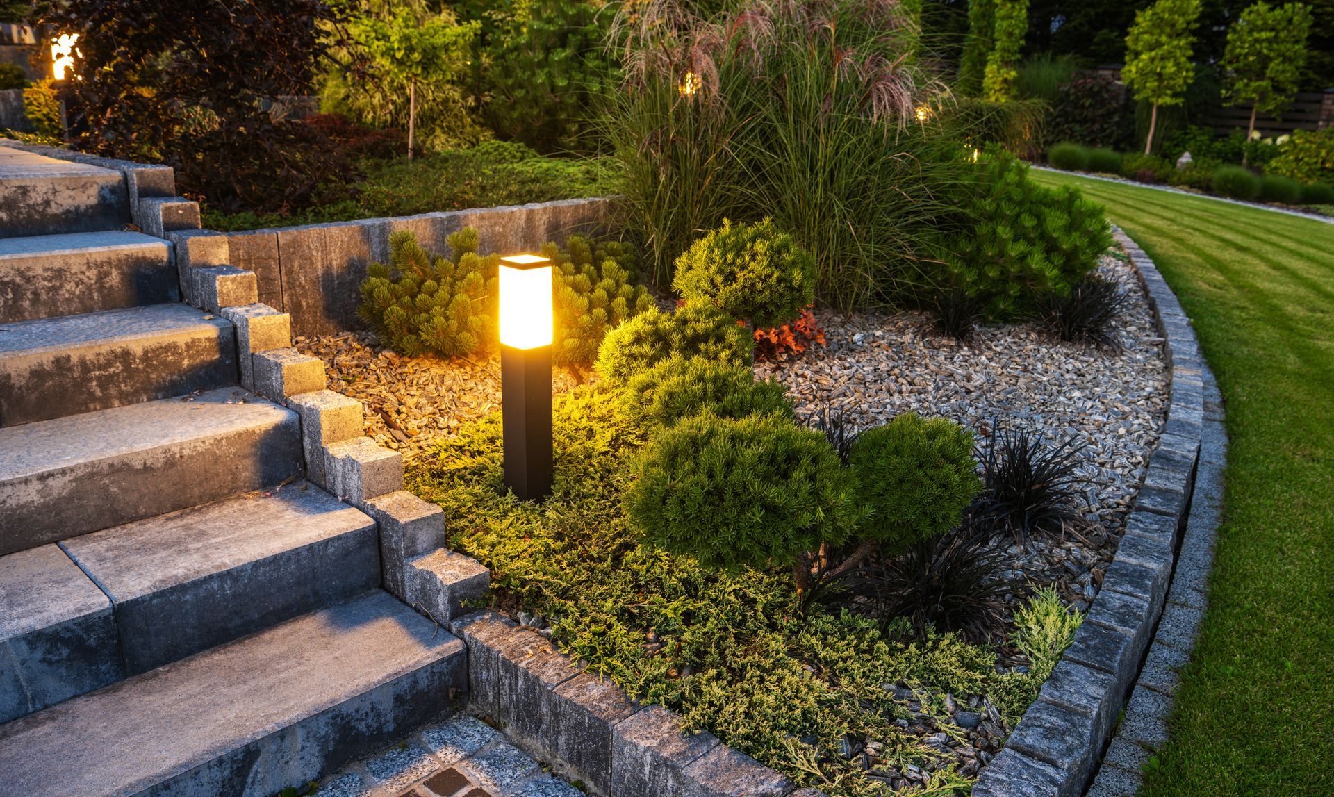 Lit stone steps beside a landscaped garden with a glowing path light at dusk.
