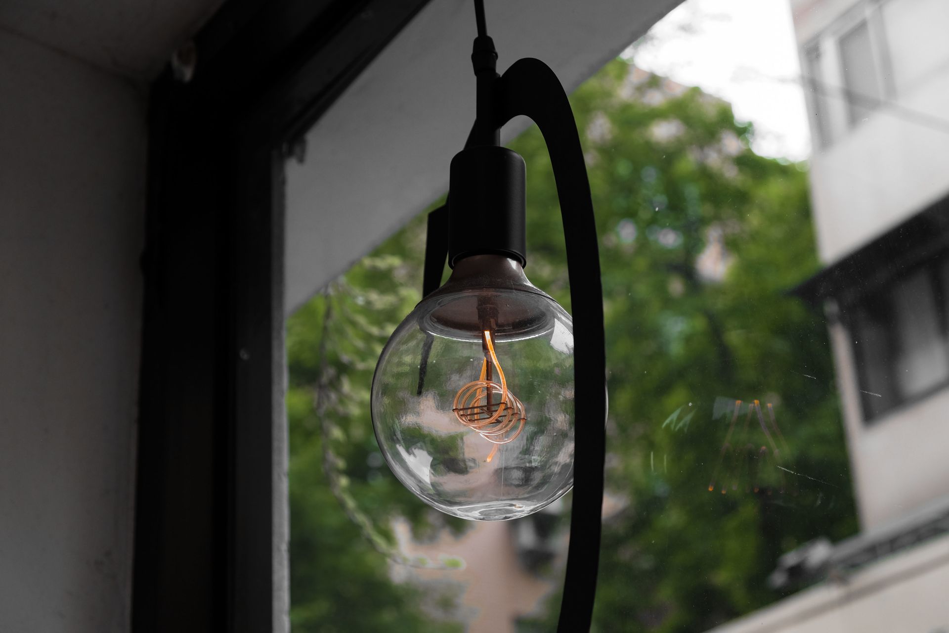 Hanging clear light bulb by a window, with blurred green trees outside and a building in the background