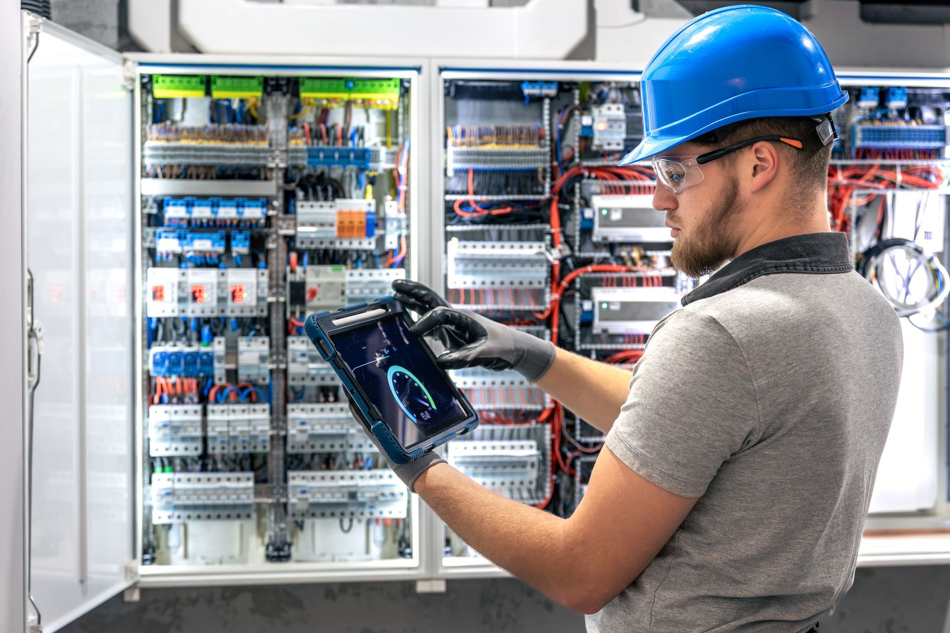 Technician in blue hard hat inspects electrical control panels with a tablet in a utility room