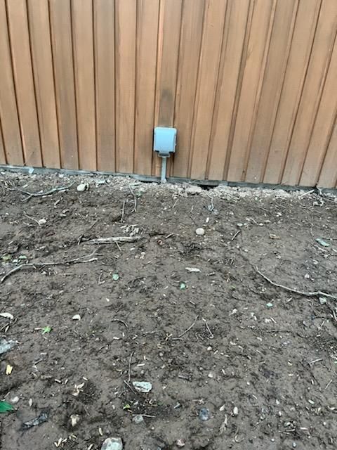 A power outlet is sitting in the dirt next to a wooden fence.