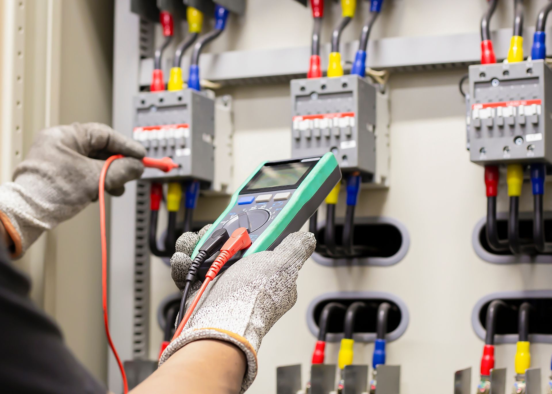 Electrician using a multimeter to test electrical panel wiring, wearing gloves.