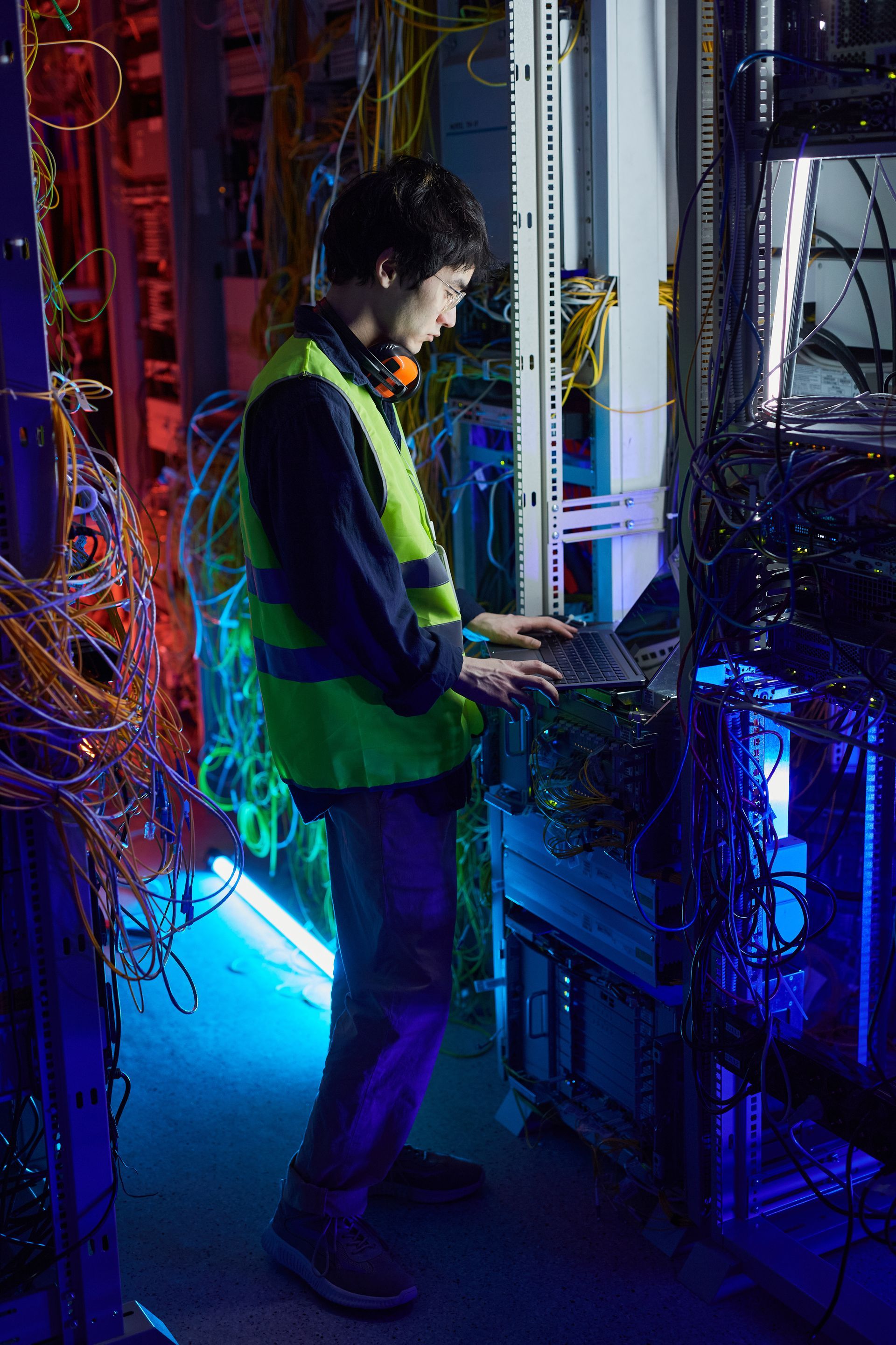 A man in a green vest is working on a laptop in a server room.