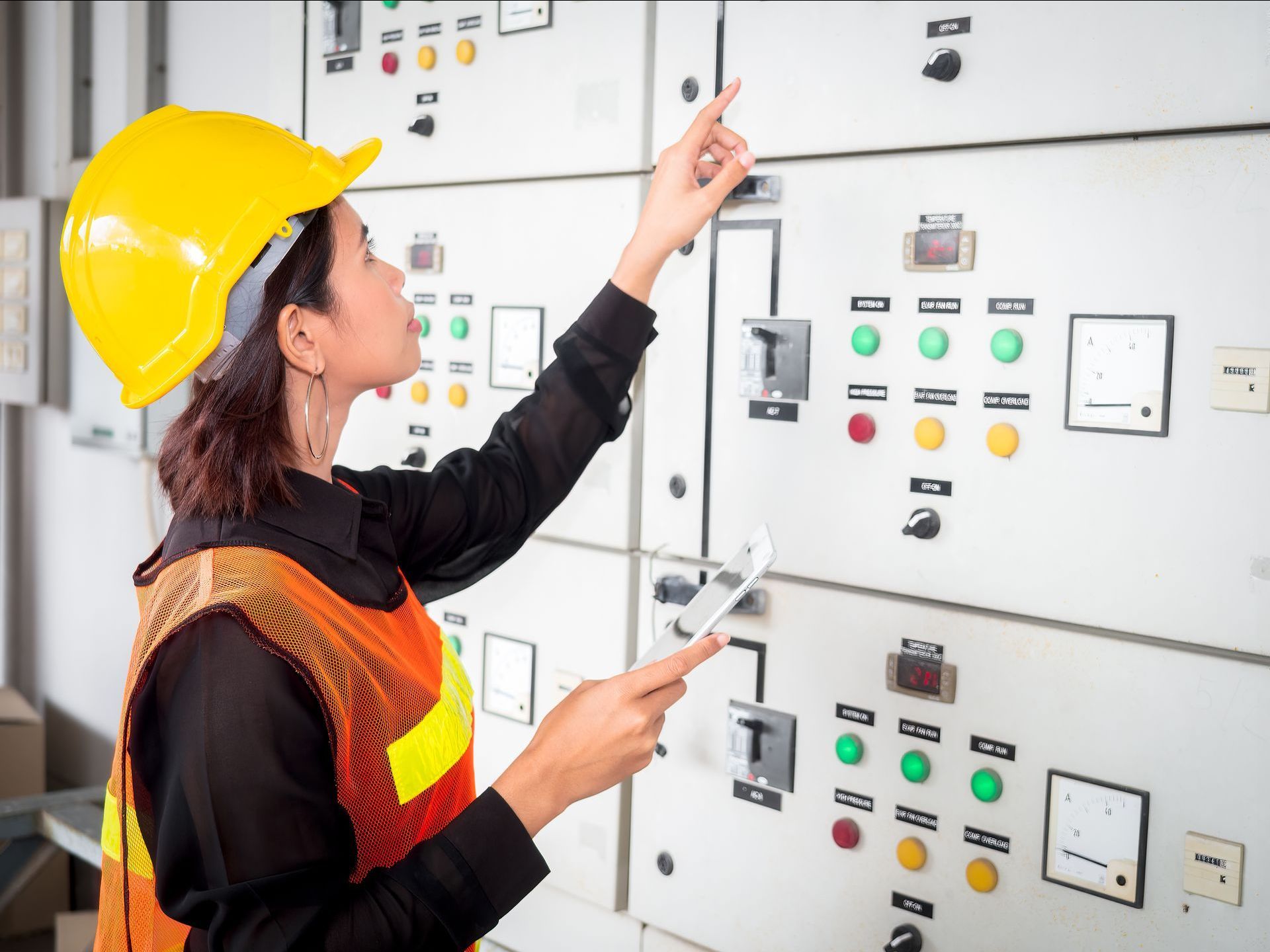 A woman wearing a hard hat and safety vest is working on a control panel.