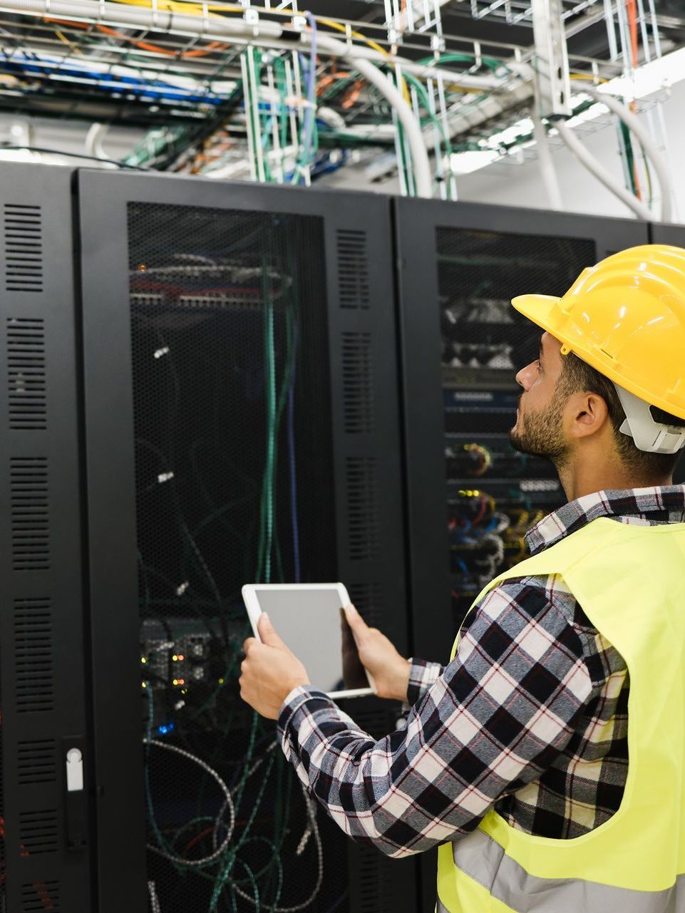 A man wearing a hard hat and safety vest is using a tablet in a server room.