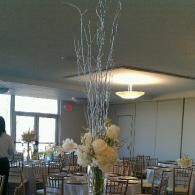 Tall floral centerpiece on a banquet table in a softly lit room with tables and chairs.
