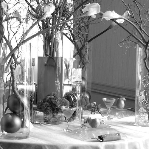 A dining table with vases, branches, fruit, flowers, and candleholders near a bright window.