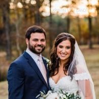 Smiling bride and groom in formal attire outdoors, with trees and warm sunset light.
