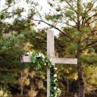 Wooden cross decorated with white flowers against a backdrop of green trees