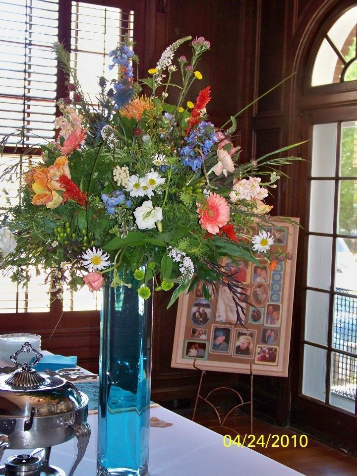 Tall blue vase with a colorful flower arrangement on a table near a window in a wood-paneled room