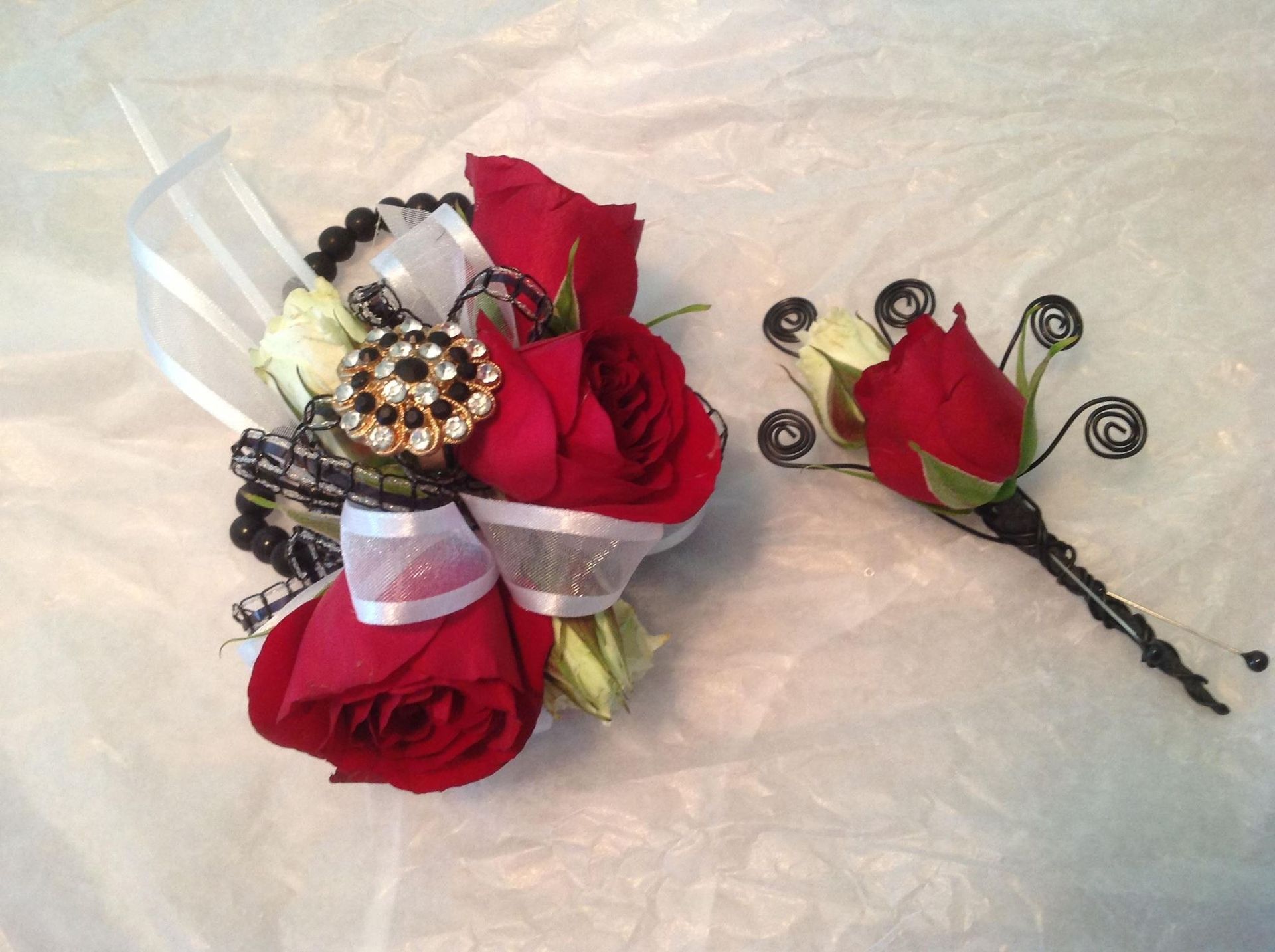 Red rose bouquet with white ribbon and jeweled accents beside a matching rose boutonniere.