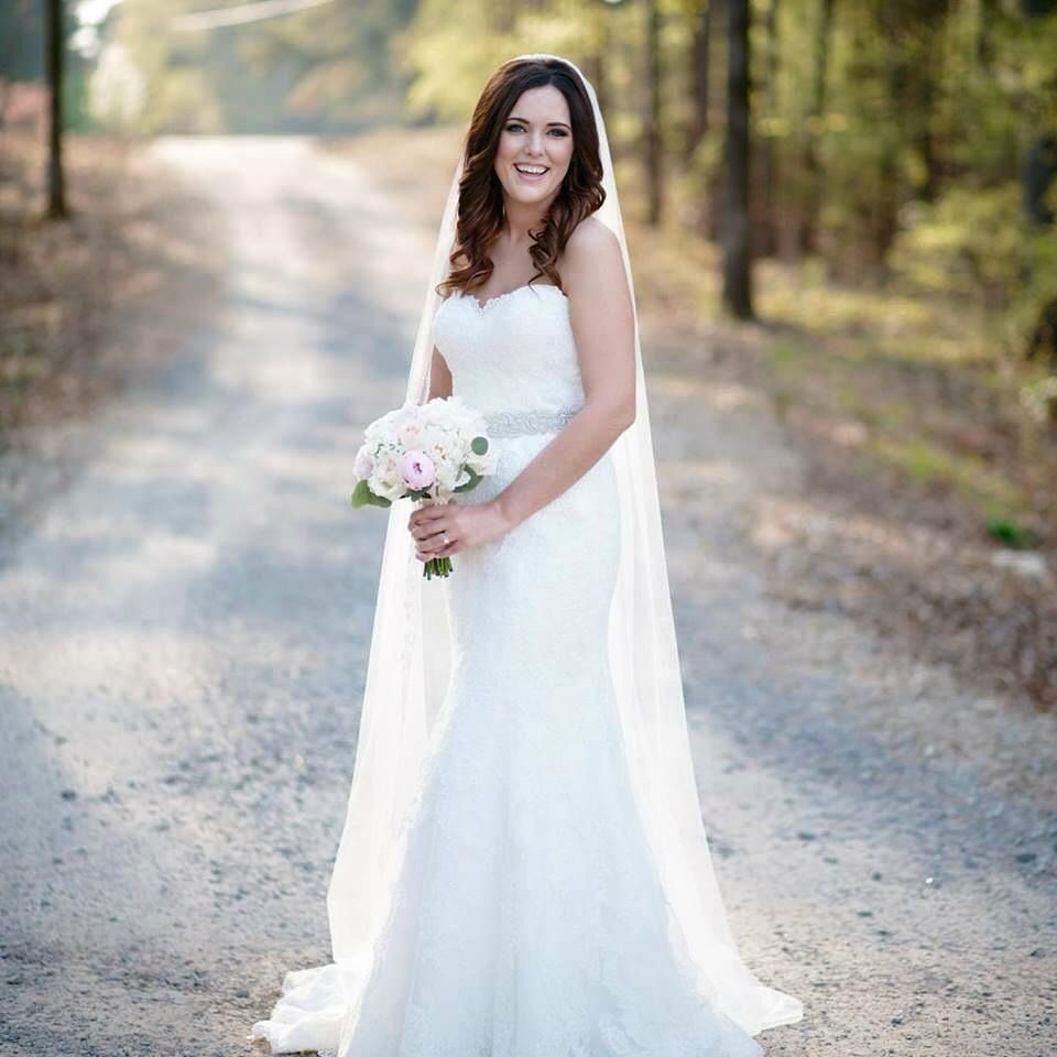 Bride in a white dress holding a bouquet, standing on a sunlit gravel path outdoors
