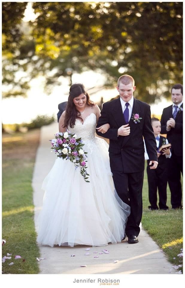 Bride and groom walking down a sunlit path, bride holding a bouquet, with wedding party behind them