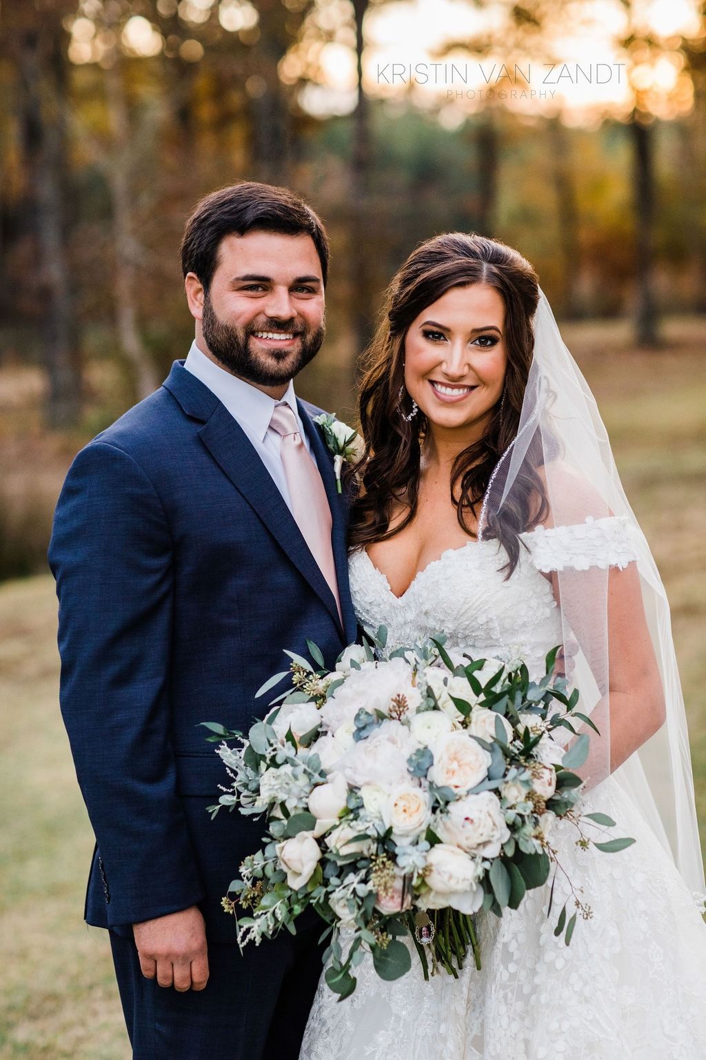 Bride and groom in wedding attire, smiling outdoors with a bouquet and autumn trees behind them