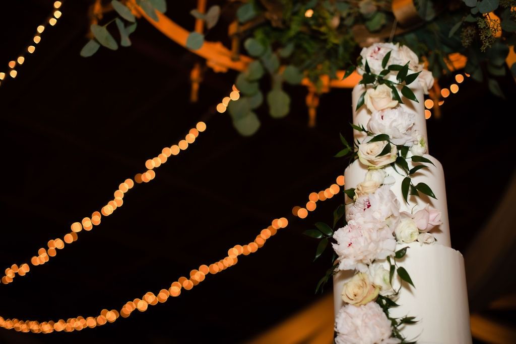 White wedding cake with pink flowers and greenery, lit by warm string lights at night