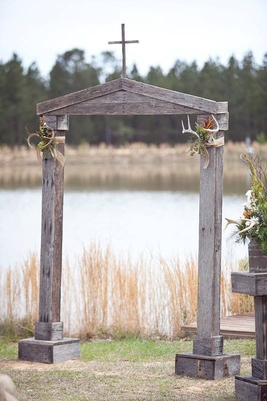 Wooden wedding arch with cross by a lakeside, decorated with flowers and flanked by tall grass