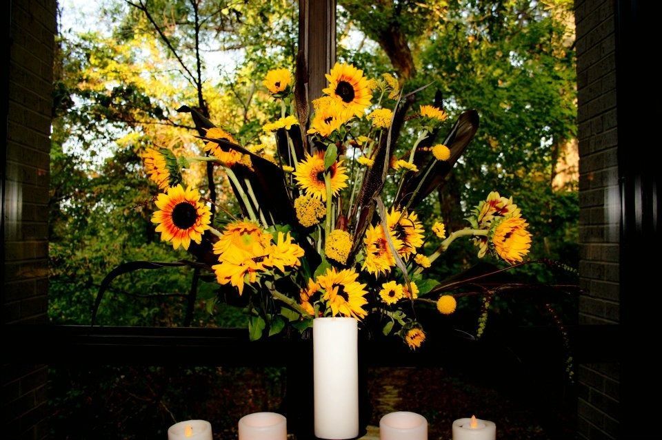 Floral arrangement with sunflowers and yellow daisies in a white vase, with candles below, in a dark setting