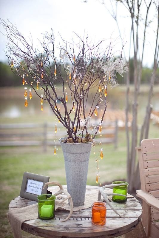 Outdoor table with a tall vase of bare branches and hanging lights, surrounded by green and orange glass candles.
