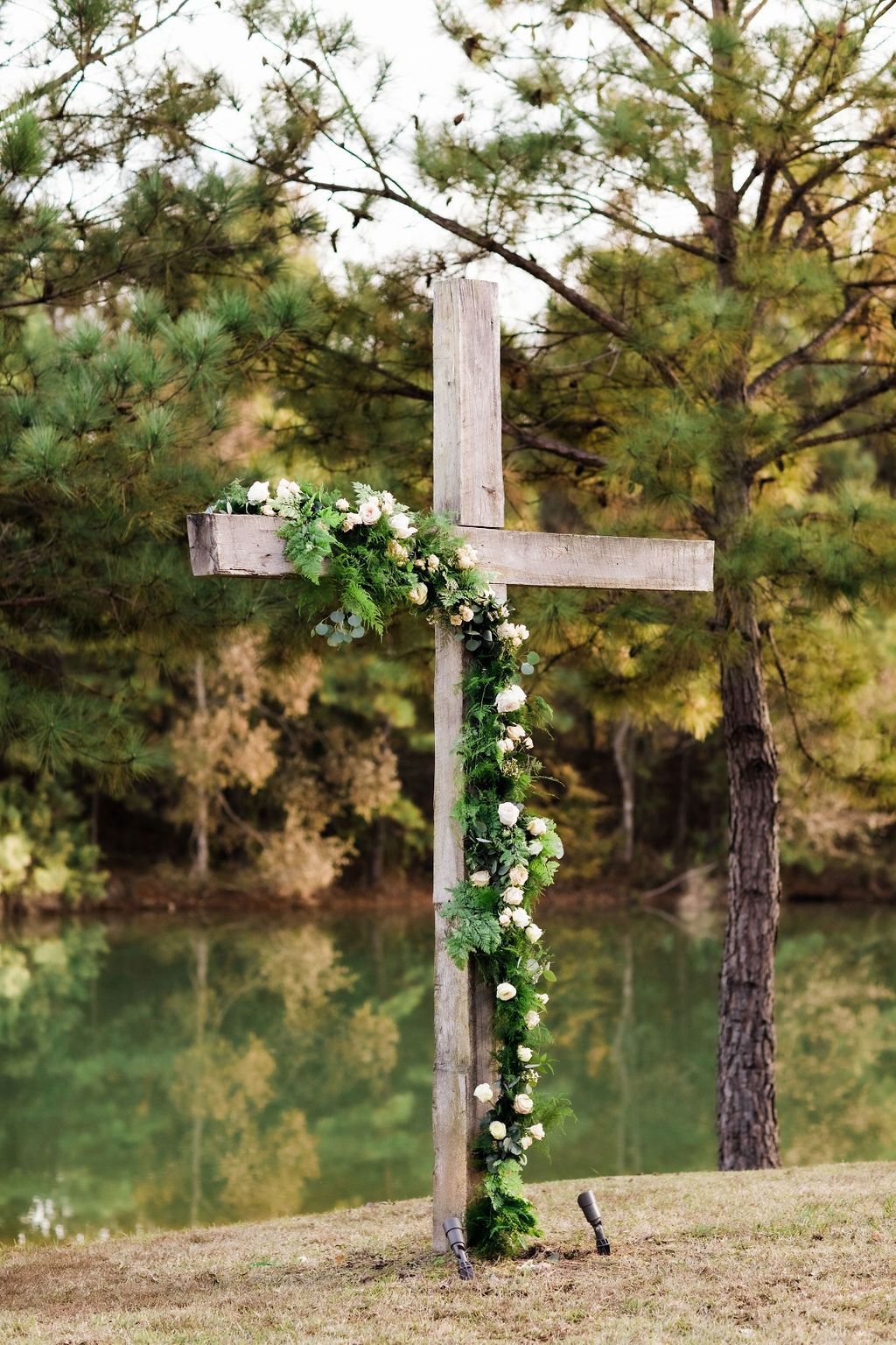 Wooden cross adorned with white flowers beside a calm lake and trees