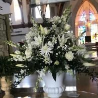 Large white floral arrangement in a vase inside a church, with stained-glass windows in the background