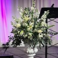 White floral arrangement in a vase on stage beside a music stand, lit with purple curtains