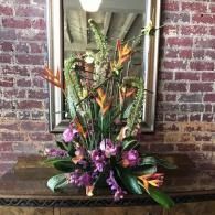 Tropical flower arrangement with purple and orange blooms on a wooden table against a brick wall and mirror