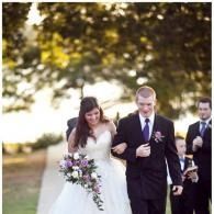 Bride and two men in dark suits walking outdoors, bride holding a bouquet under trees