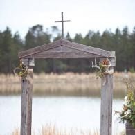 Wooden wedding arch with cross beside a lake, decorated with flowers and greenery.