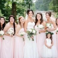 Bridesmaids in blush dresses holding white bouquets, standing with a bride and flower girl outdoors