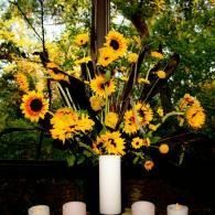 Sunflower bouquet in a white vase with candles on a table, set against a dark leafy background
