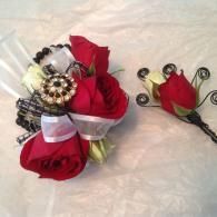 Red rose corsage with white ribbon and jeweled center beside a matching rose boutonniere on white fabric