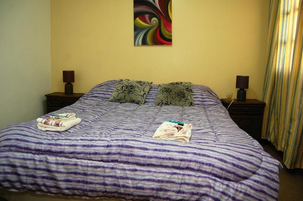 Dormitorio con colcha morada, dos lámparas de noche y un colorido cuadro en la pared encima de la cama.