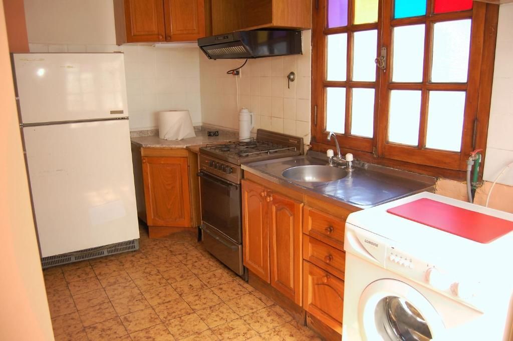 Cocina pequeña con nevera blanca, armarios de madera, fogones, fregadero y lavadora junto a la ventana.
