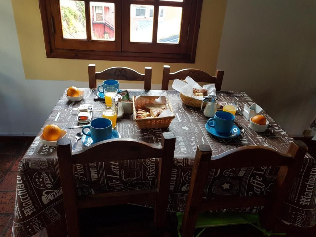 Prepara la mesa del desayuno junto a la ventana con tazas, platos, pan y mermelada sobre manteles estampados.
